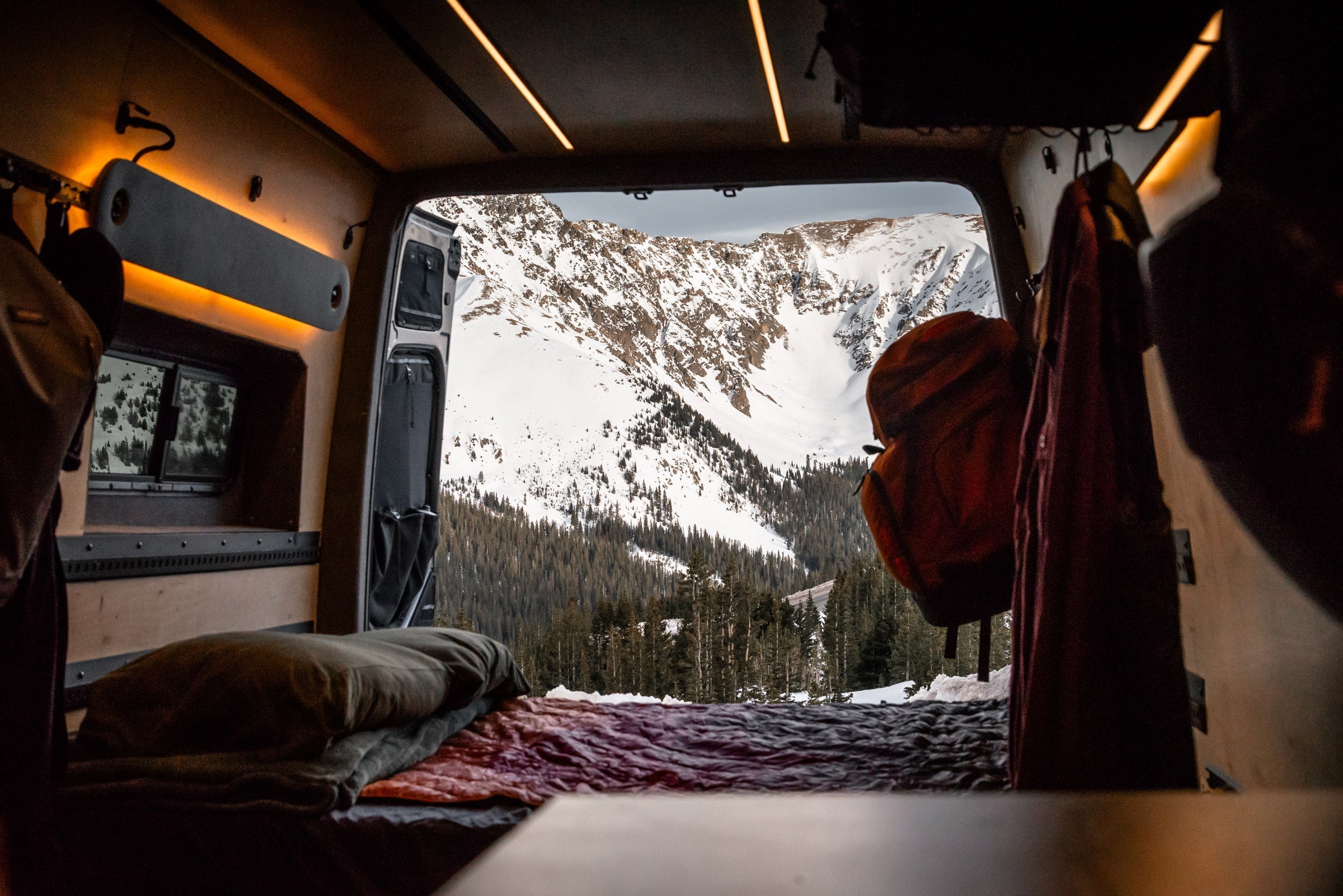 View from inside a camper van looking out at snow-covered mountains and pine trees, with bedding and backpacks visible in the foreground.
