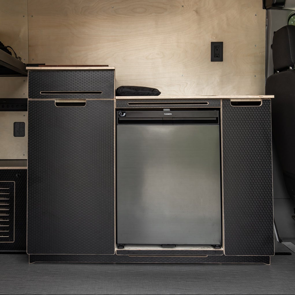 Compact kitchen area inside a vehicle with cabinets and a refrigerator.