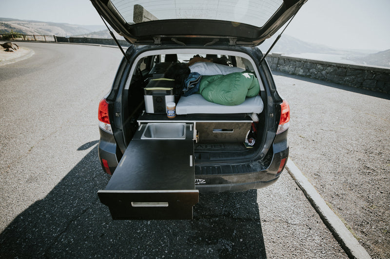 Car trunk with the Trekhaus SUV Camper System, showing off a bed setup and drawer extended, on a road.