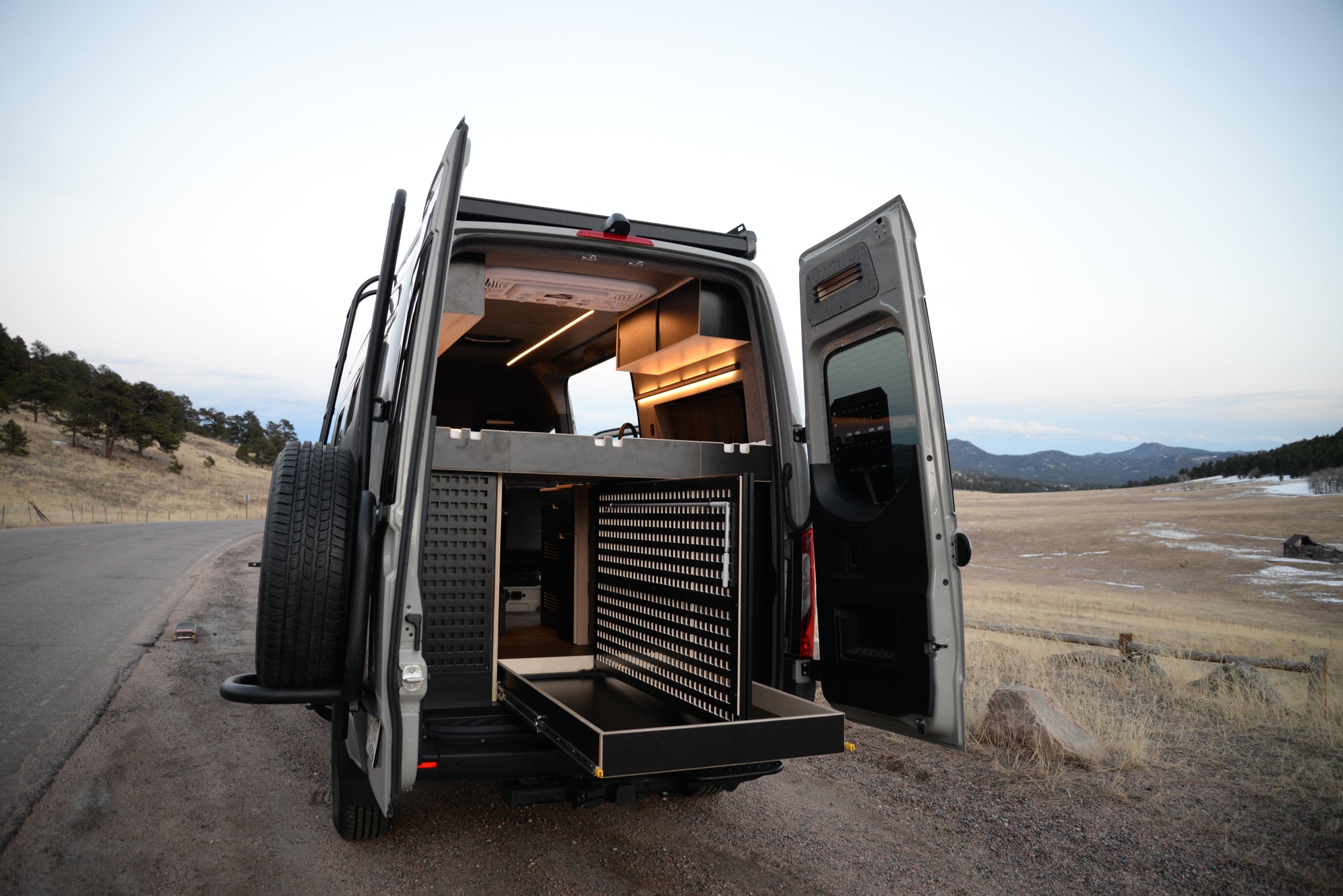 Camper van with open rear doors showing off the Venturhaus Modular Van System. Landscape is scenic with trees, mountains and snow in some areas